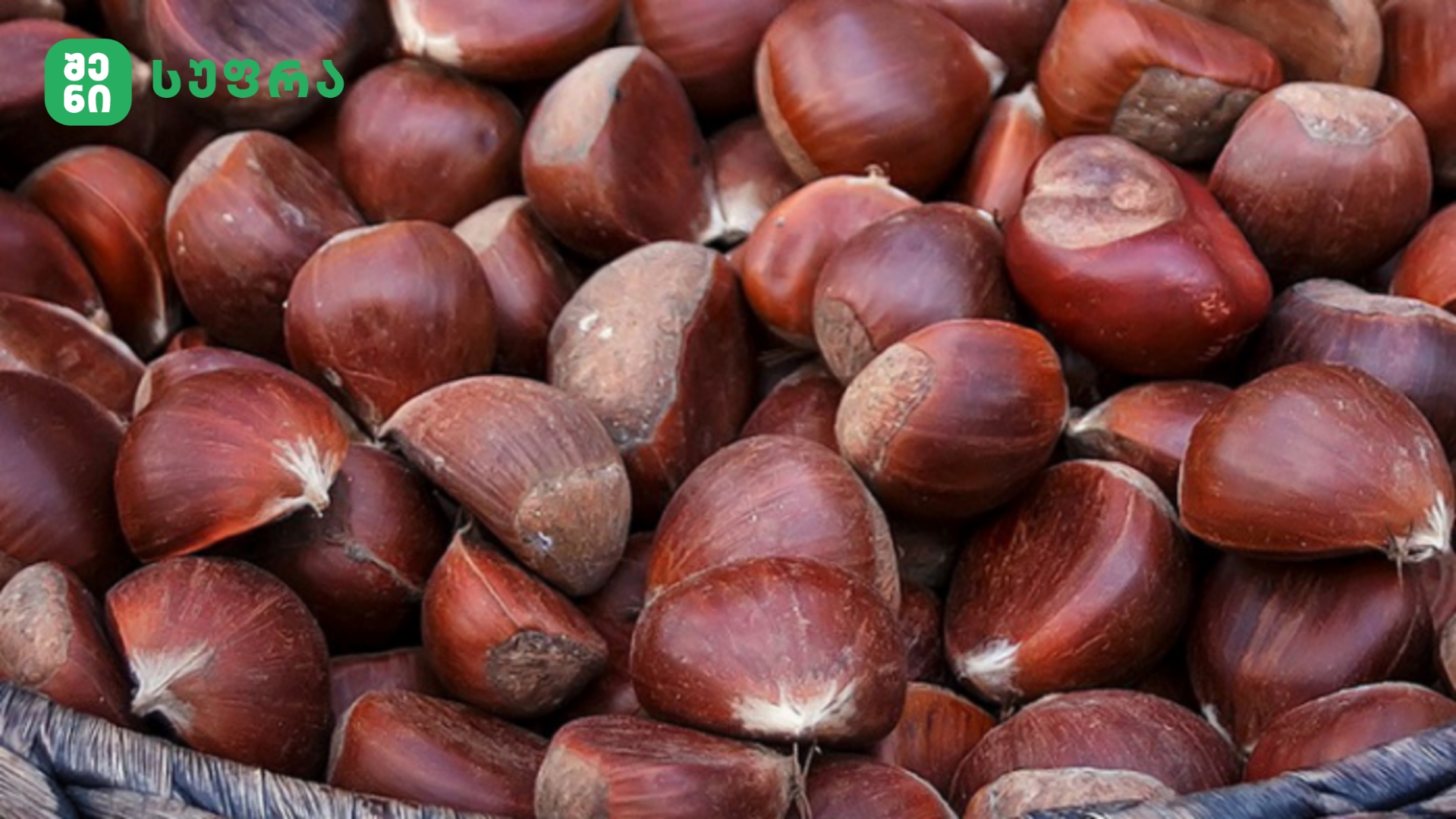 Close-up of glossy chestnuts piled in a basket with a green logo in the corner.