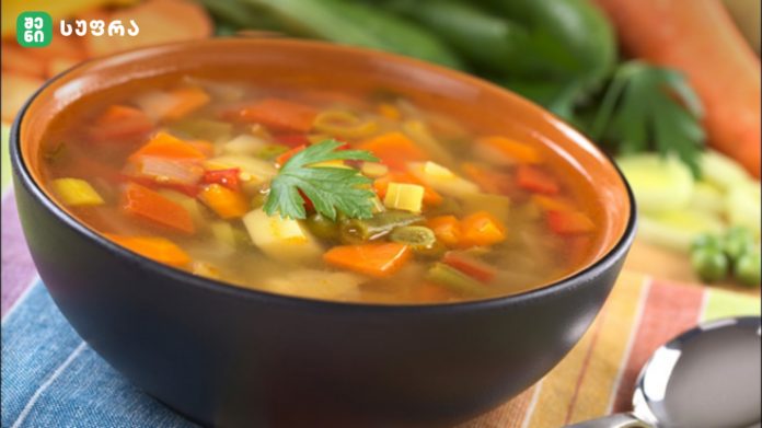 Bowl of vegetable soup with carrots, potatoes, and herbs, garnished with parsley on a colorful table, logo in the corner (top-left).