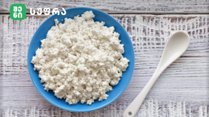 Blue bowl filled with cottage cheese on a whitewashed wooden table, with a white spoon nearby and a logo in the top-left corner.