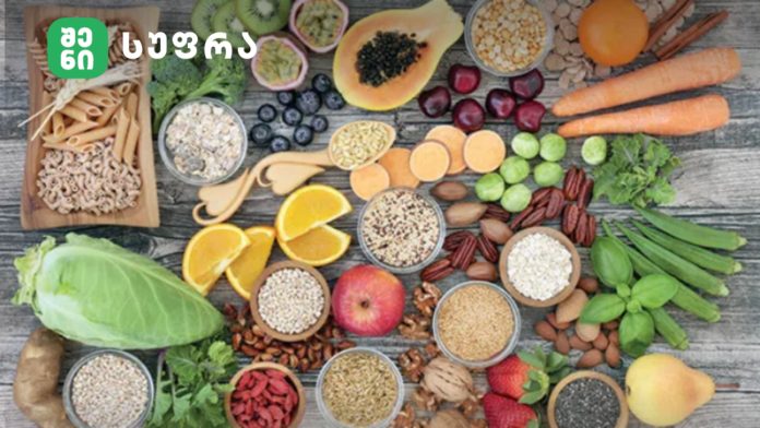 Assorted healthy foods spread on a weathered wooden table, with a green logo in the top-left corner (fruits, vegetables, grains, nuts, and seeds).