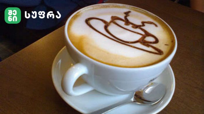Latte with heart-shaped chocolate drizzle in a white cup on a saucer, on a wooden table, with a green logo in the top-left corner.