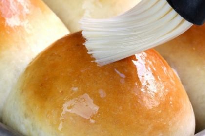 Close-up of golden-brown dinner rolls being brushed with glaze using a pastry brush.