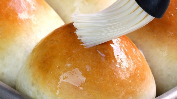 Close-up of golden-brown dinner rolls being brushed with glaze using a pastry brush.