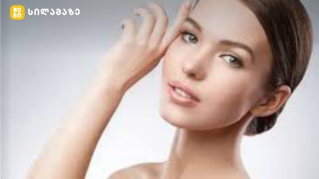 Close-up portrait of a woman with light makeup, touching her temple with her right hand; studio lighting, watermark/logo in the top-left corner.