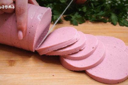 Thin rounds of pink deli sausage being sliced on a wooden cutting board with fresh herbs in the background.