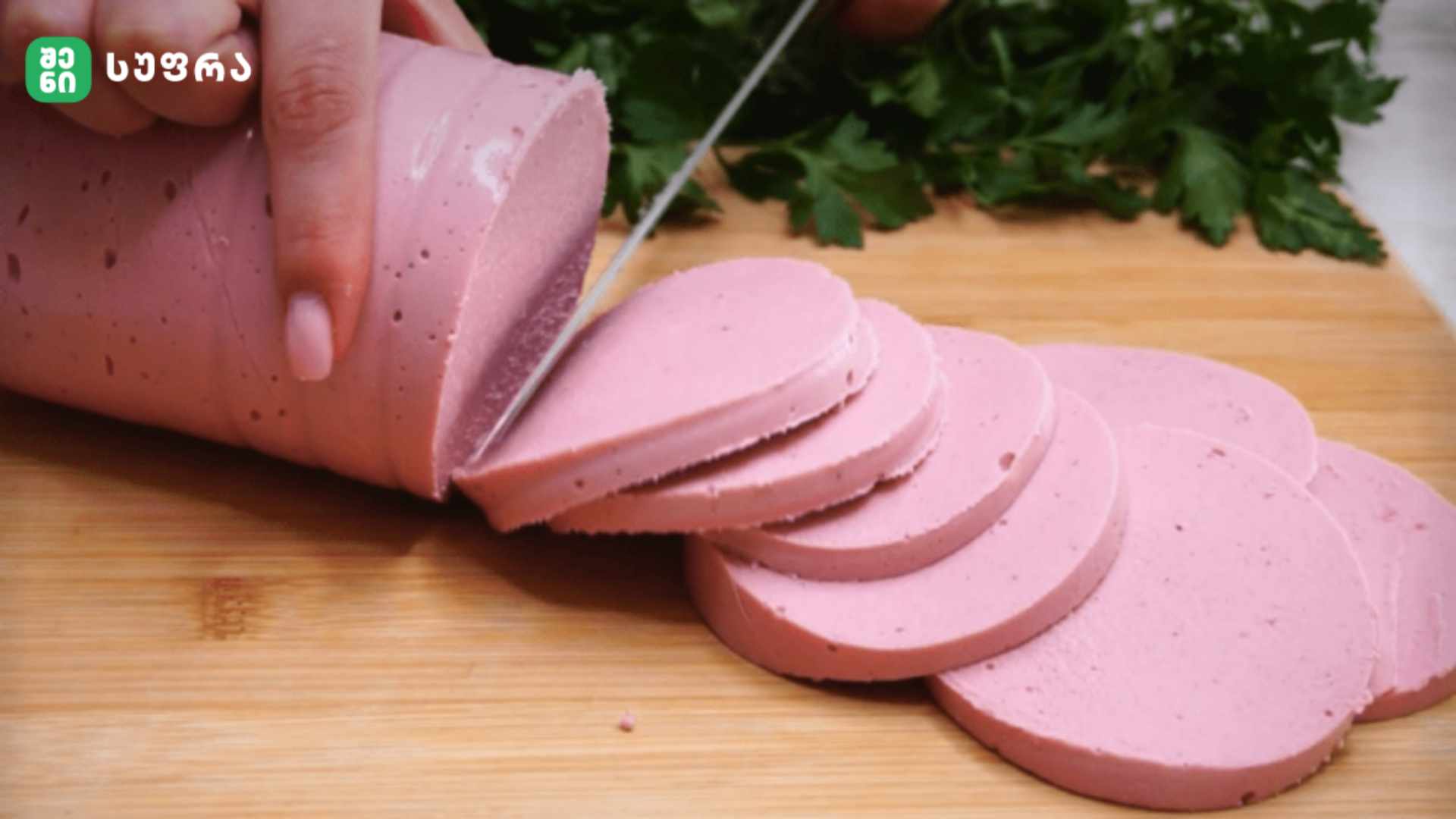 Thin rounds of pink deli sausage being sliced on a wooden cutting board with fresh herbs in the background.