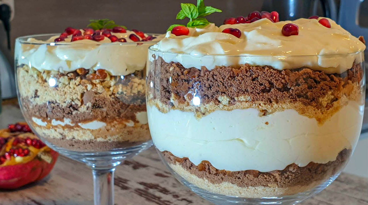 Layered dessert in a glass cup with cream, crumb, and chocolate layers, topped with pomegranate seeds and a mint leaf garnish.